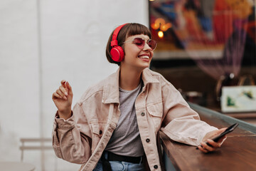 Joyful teen girl with brunette hair listening to music outside. Short-haired lady in red...
