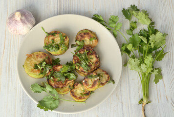 Fried zucchini lie on a light plate, which stands on a light table, next to herbs an