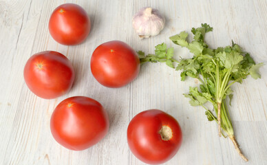Five red large tomatoes are next to a bunch of greens and a head of garlic on a light background
