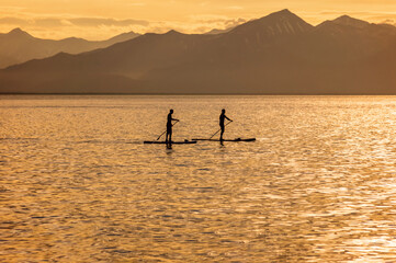 Silhouettes of two men standing on sup board against the mountains on the ocean at sunset.