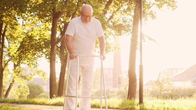 Disabled old man is walking with a walker. Handicapped patient in the park. Assistance, rehabilitation and health care. Golden hour sunset.