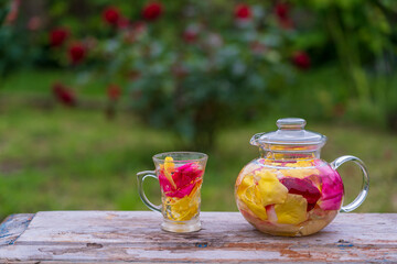 Beautiful and fragrant tea from yellow, pink and red petals roses in glass teapot and mug in the summer garden on wooden table. Close up