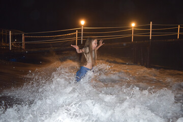 An eight-year-old girl stands in the spray from the sea wave and waves her hands in surprise on a...