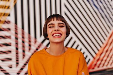 Joyful woman with brunette hair smiling widely on striped background. Trendy short-haired lady in...