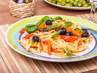 Classic vegetarian pasta - linguini with tomatoes, peppers and olives with basil leaves, serving on a plate on the table in a cafe, close-up