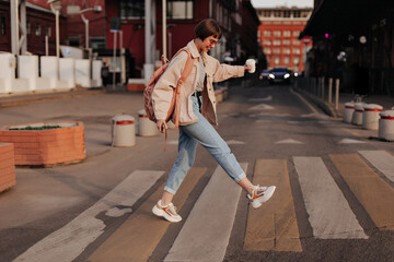 Happy lady walking at street. Brunette woman in light jacket and jeans with brown backpack smiling...