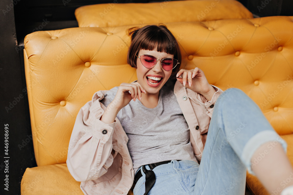 Wall mural excited woman with short hair sitting on yellow couch inside. modern girl in pink glasses, jeans and