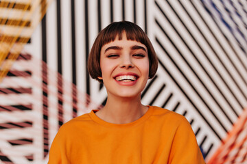 Happy woman with short hairstyle smiling widely on striped background. Good-humored lady in orange...