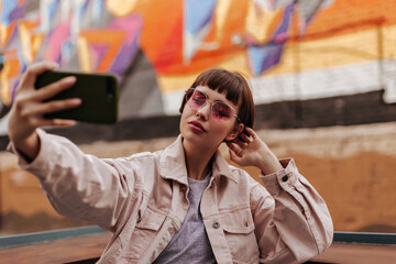 Beautiful teen girl taking photo outside. Short-haired lady with beige outfit and pink sunglasees...
