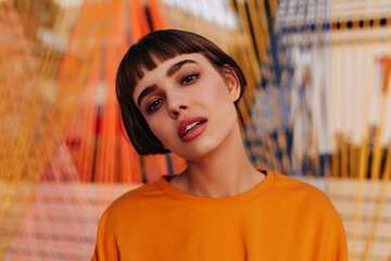 Beautiful girl with brunette hair in orange outfit posing on striped backdrop. Stylish woman in...