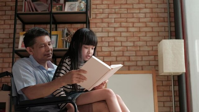 Granddaughter sat on the old Asian grandfather's lap, disabled and retired in a wheelchair in the house, while happily reading book together during the vacation. Love for a relationship lifestyle.