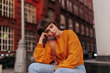 Short-haired woman in orange sweater sits outdoors. Young hipster girl in bright sweatshirt and...