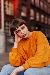 Thoughtful brunette short-haired woman sits outside. Attractive young girl in orange sweatshirt and jeans looks into camera.