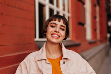 Attractive short-haired woman in pink denim jacket and orange sweater smiles outside. Happy girl...