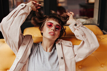 Fashionable teen girl in bright sunglasses posing on yellow couch inside. Short-haired woman in...