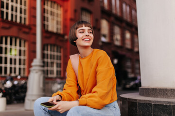 Good-humored woman sits outside. Short-haired girl in orange sweater holds smartphone and smiles...