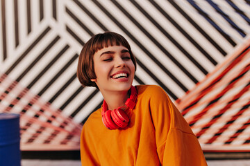 Joyful lady in orange outfit sincerely smiling on striped backdrop. Brunette teen girl with bright...