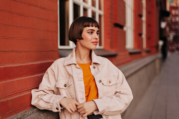 Cool woman in pink denim jacket and orange sweatshirt poses outside near brick building. Attractive girl walks outdoors. - obrazy, fototapety, plakaty