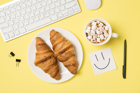 Top view photo of workplace white keyboard mouse binder clips pen sticker note with drawn smiling face cup of drink with marshmallow and plate with two croissants on isolated light yellow background