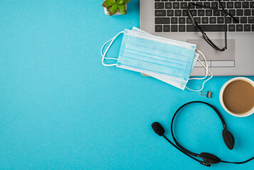 Top view photo of glasses two medical masks on laptop binder clip headphones flowerpot and cup of drink on isolated pastel blue background with empty space