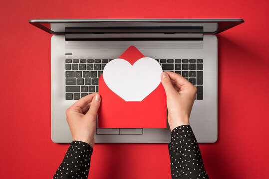 Overhead Photo Of Grey Laptop And Hands Holding A Red Envelope With White Paper Heart Inside Isolated On The Red Backdrop