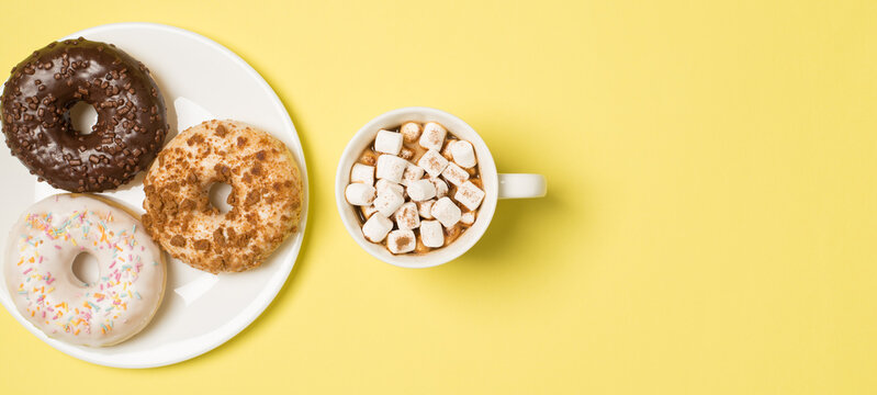 Top view photo of cup of drink with marshmallow and plate with three different color fresh donuts on isolated pastel yellow background with empty space
