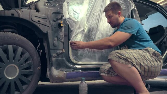 A painter worker who seals the holes of a car and other body elements with paint tape so as not to fill it with paint during painting in a car repair shop. Pasting with a film.