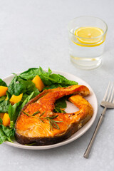 Fried salmon steak with spinach and yellow tomatoes in a plate on a gray background, a glass of water with lemon, a hearty lunch with healthy red fish