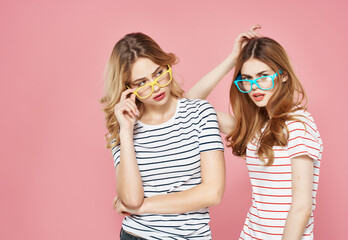 two girlfriends hugging in striped t-shirts multicolored glasses pink background
