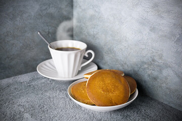 Pancakes in dish and coffee with steam in porcelain cup on gray surface, white tableware with pastries and hot drink.
