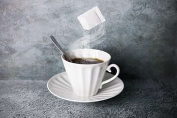 Levitating sachet of pouring sugar over white porcelain hot coffee cup with steam, spoon and saucer...