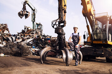 Portrait of junkyard worker with hardhat standing next to hydraulic industrial machine with claw attachment used for lifting scrap metal parts. - obrazy, fototapety, plakaty