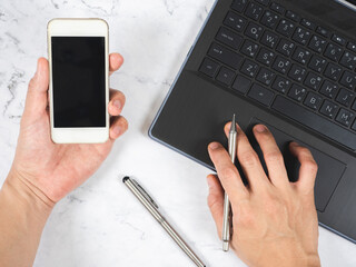 Top view workspace hand holding mobile phone with silver pen and laptop white marble desk