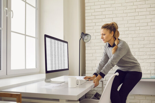 Financial accountant working with online bookkeeping spreadsheets. Serious young woman sitting on edge of office desk and looking at screen of desktop computer standing on table with cup of coffee