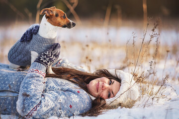 Young woman in winter jacket lying down at snow covered ground, holding her Jack Russell terrier...