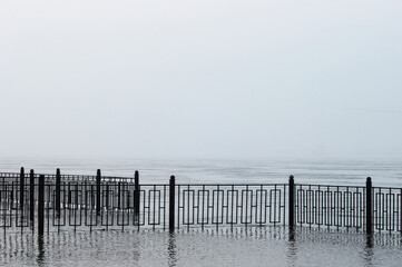 Fence of the city embankment during flooding with water. Very heavy fog. Colorless early morning before sunrise.