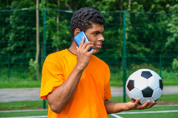 Portrait of young Brazilian man holding soccer ball and use smartphone while standing on sports court outdoors against soccer goal