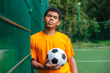 Portrait of young Brazilian man holding soccer ball while standing against fence in sports court outdoors, copy space