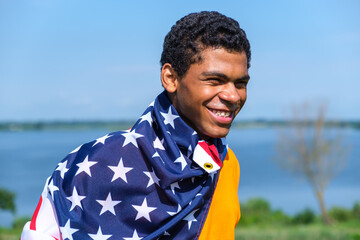 Young smiling African American man proudly holding American flag covered on his shoulders against blue sky in summer