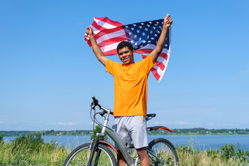 Handsome young afro American man holding and waving USA flag and standing with bike on summer meadow against blue sky background in morning