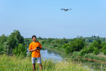 Full length portrait of young smiling African American man operating drone with controller in hands on the meadow near the river. 