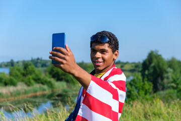 Cheerful smiling African American young man in sunglasses and covered with American flag taking selfie outdoors.