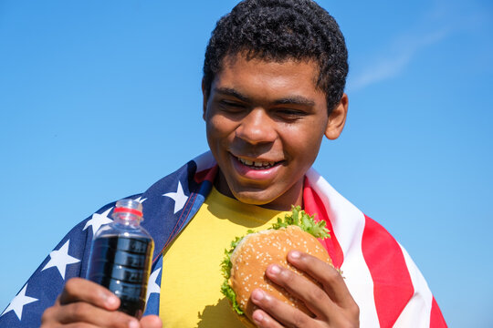 Hungry African American man enjoying the taste of hamburger and cola drink while sitting wrapped in american flag outdoor in summer close-up. Concept of love to junk food diet 
