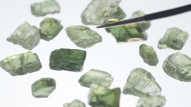Translucent uncut emeralds lie on a bright white table. The tweezers lift one stone up. Geologist examines rocks. Close-up, 4K UHD.