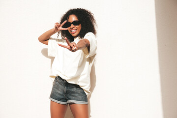 Beautiful black woman with afro curls hairstyle.Smiling hipster model in white t-shirt. Sexy carefree female posing in the street near white wall in sunglasses. Cheerful and happy.Shows peace sign