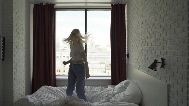 Young funny woman cheerfully jumps on the big bed in the bedroom. She enjoys life. Cozy apartment, female joy. Slow motion, HD.