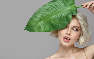 Portrait of blonde woman and green leaf. Organic cosmetics concept. Gray background. - 446935200