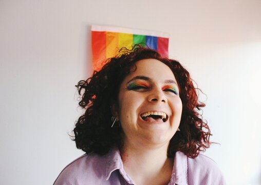 portrait of lgbtq queer lesbian young woman happy laughing & smiling happy with rainbow pride flag eye makeup and red hair curls