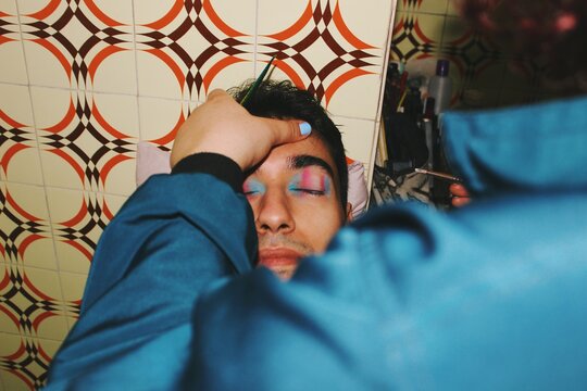 portrait face of lgbt queer young man smiling getting pink blue & white colored trans pride flag makeup from makeup artist with flash at night *3