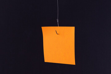 Mockup of a Blank Orange Memo Paper with Copy Space Hanging on a Fishing Hook Against the Black Background. Sticky Note Template